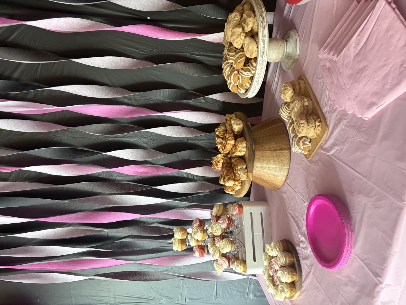 Birthday dessert table with a pink and black streamer backdrop, a cupcake tower, sticky buns on a wooden stand and iced cookies