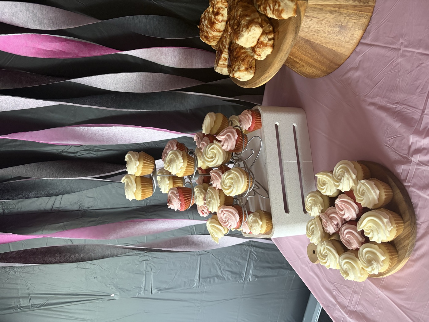 Tiered stand of pink and vanilla frosted cupcakes beside a platter of cupcakes and a wooden stand of sticky buns