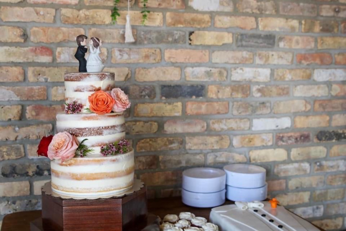 Three-tier naked wedding cake with roses against a brick wall, beside a groom’s cake and cookies